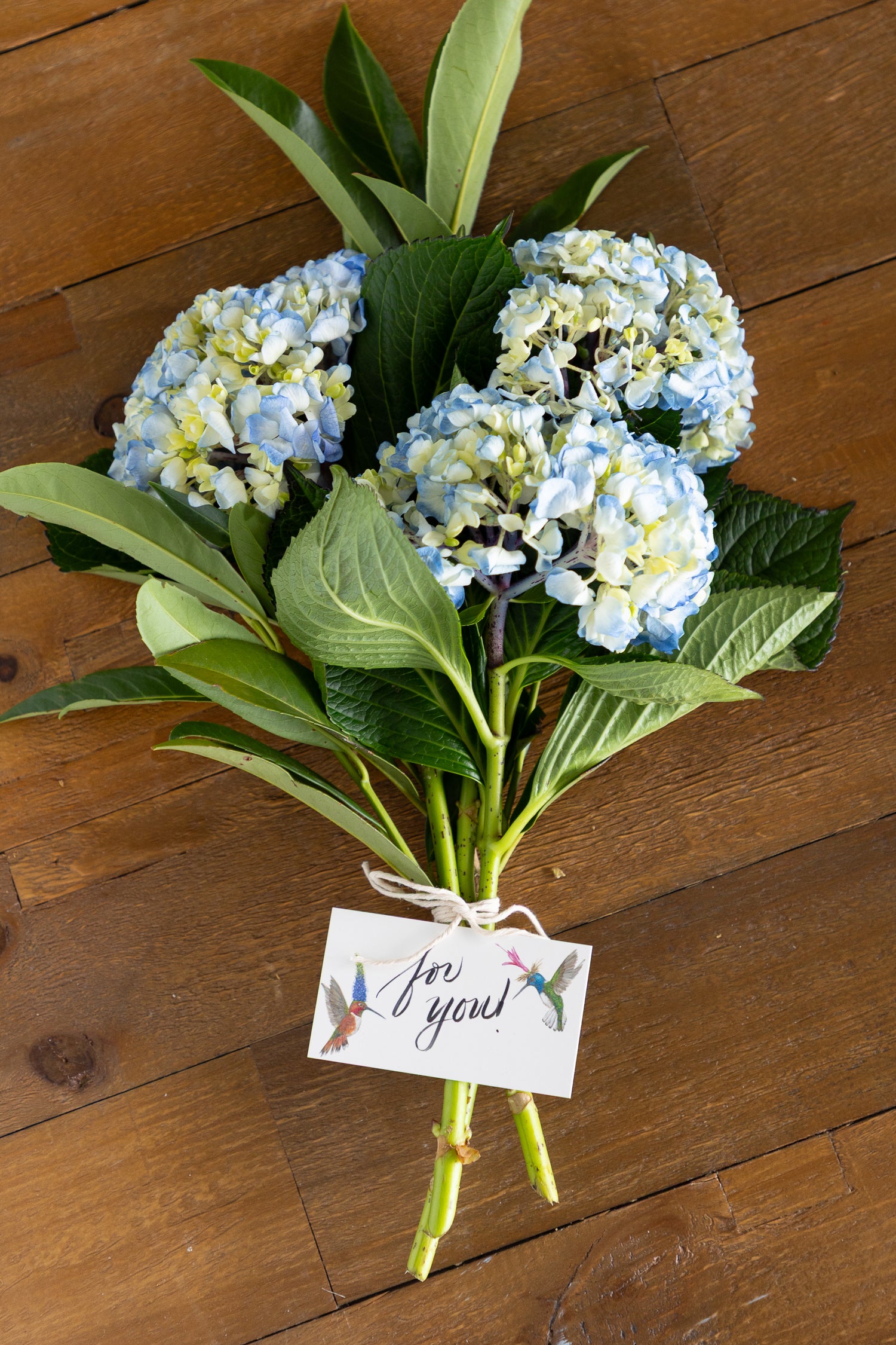 A bouquet of light blue and white hydrangeas with green leaves, tied with a string and a small card that says &