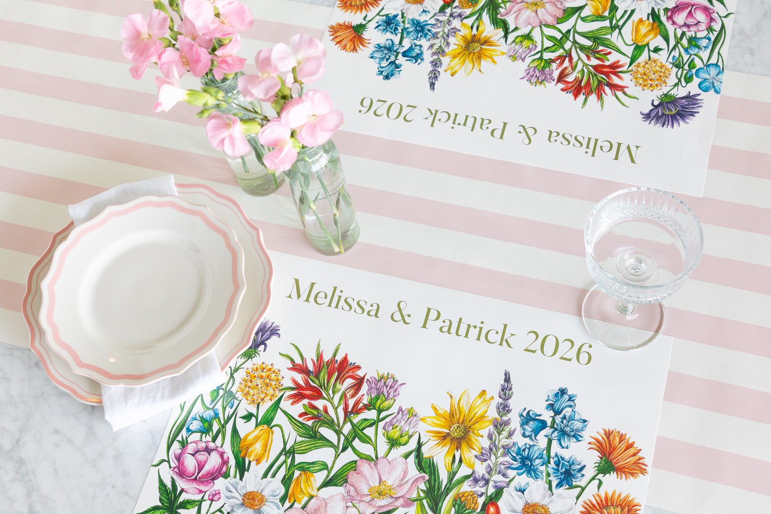 Personalized floral place mats on a pink and white striped tablecloth, styled with a white plate, a glass goblet, and pink flowers in a glass bottle.