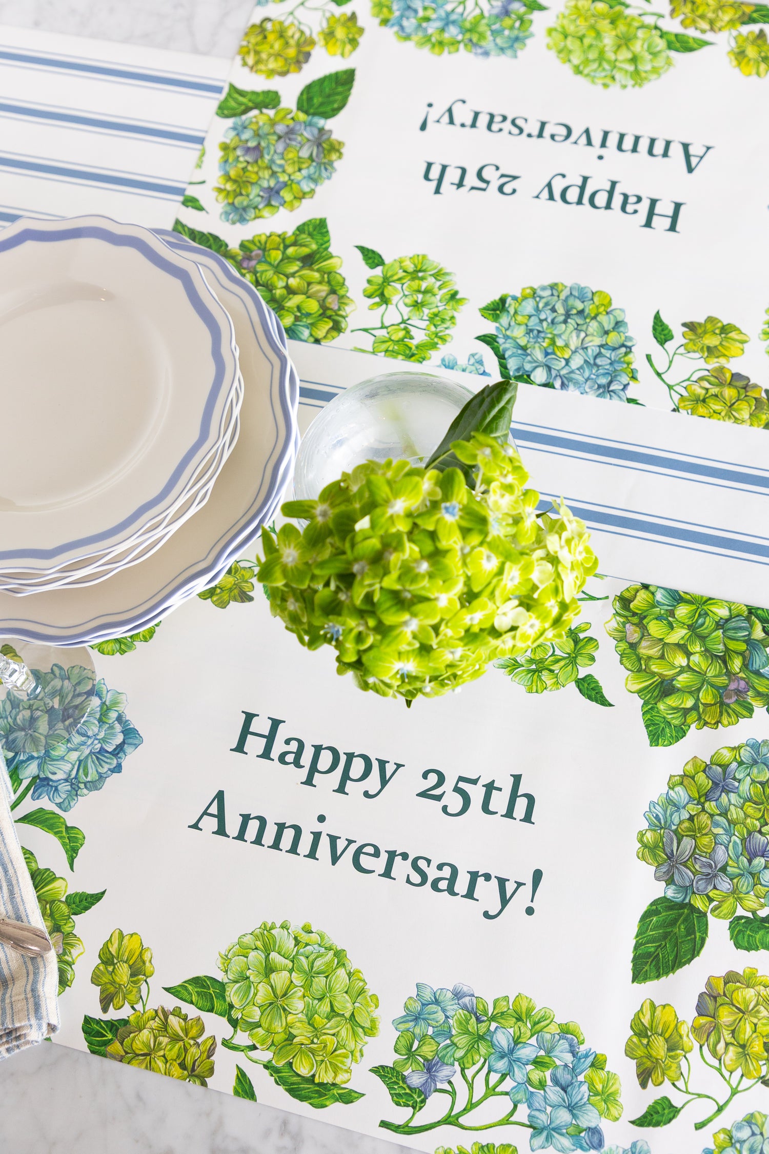 A close-up of a table setting featuring printed paper placemats with a 25th anniversary design. The placemats are decorated with watercolor-style green and blue hydrangeas and the text &