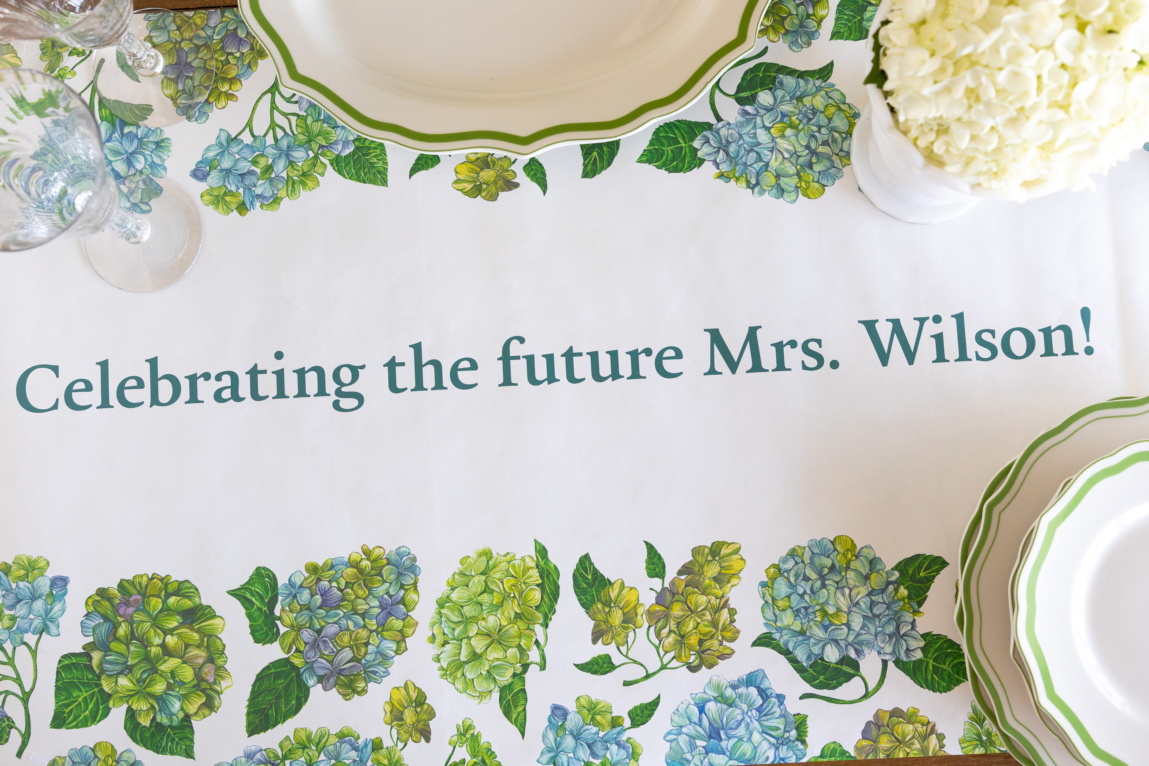 A white table runner with a green and blue floral border and the printed text &