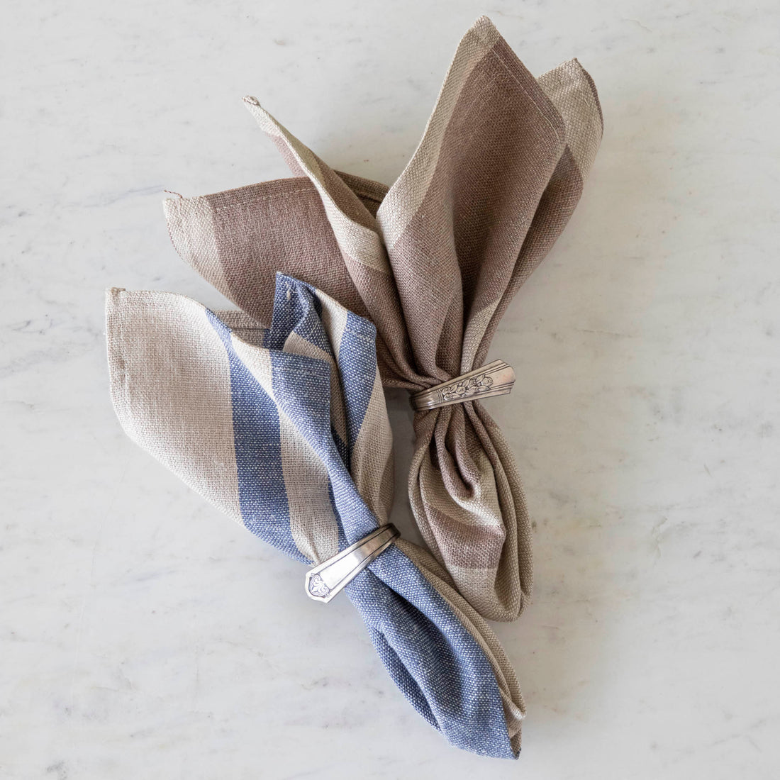 A Vintage Blue Striped Woven Linen Napkin and Dusty Rose Striped Woven Linen Napkin in silver napkin ring holders on a marble table.