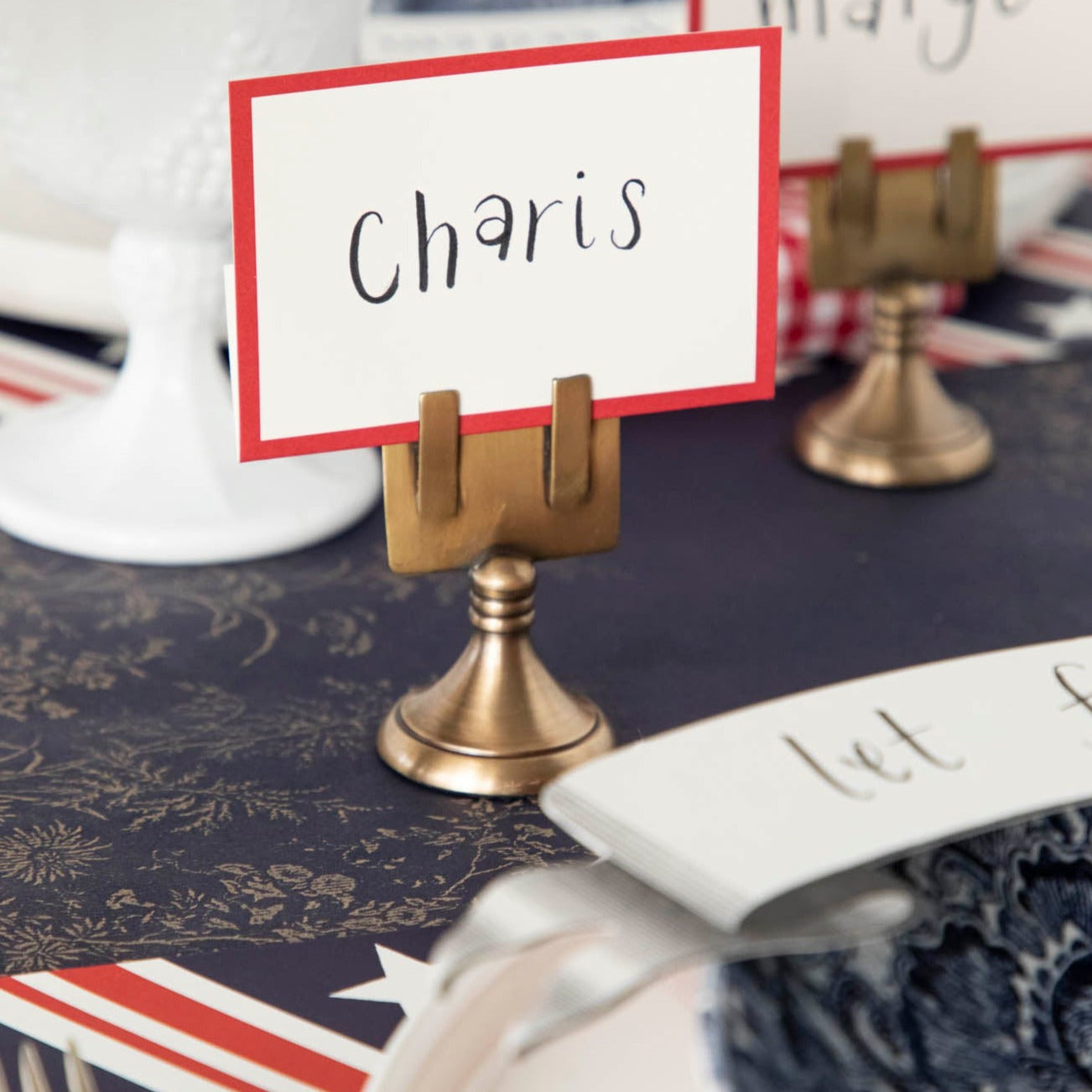Close-up of a Red Frame Place Card labeled &quot;Charis&quot; held by a brass table card holder on a nautical-themed table setting.