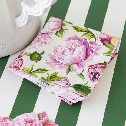 A pink Peony Napkin from Hester &amp; Cook on a green and white striped table, perfect for a party with fresh flowers.