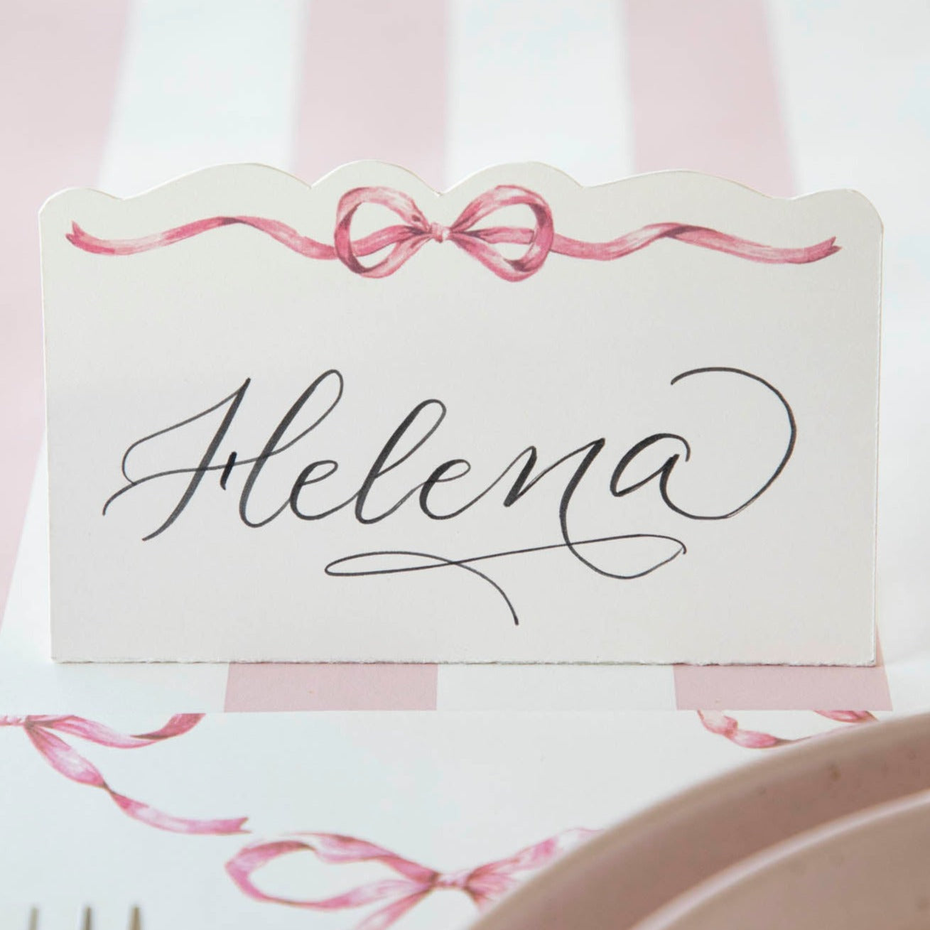 This Pink Bow Place Card, from Hester &amp; Cook, rests delicately on a table amidst dishes and exudes loveliness.