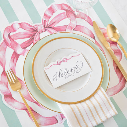 A whimsical Die-cut Pink Bow Placemat place setting with a die-cut pink bow accent, including a plate, gold spoon, and fork by Hester &amp; Cook.
