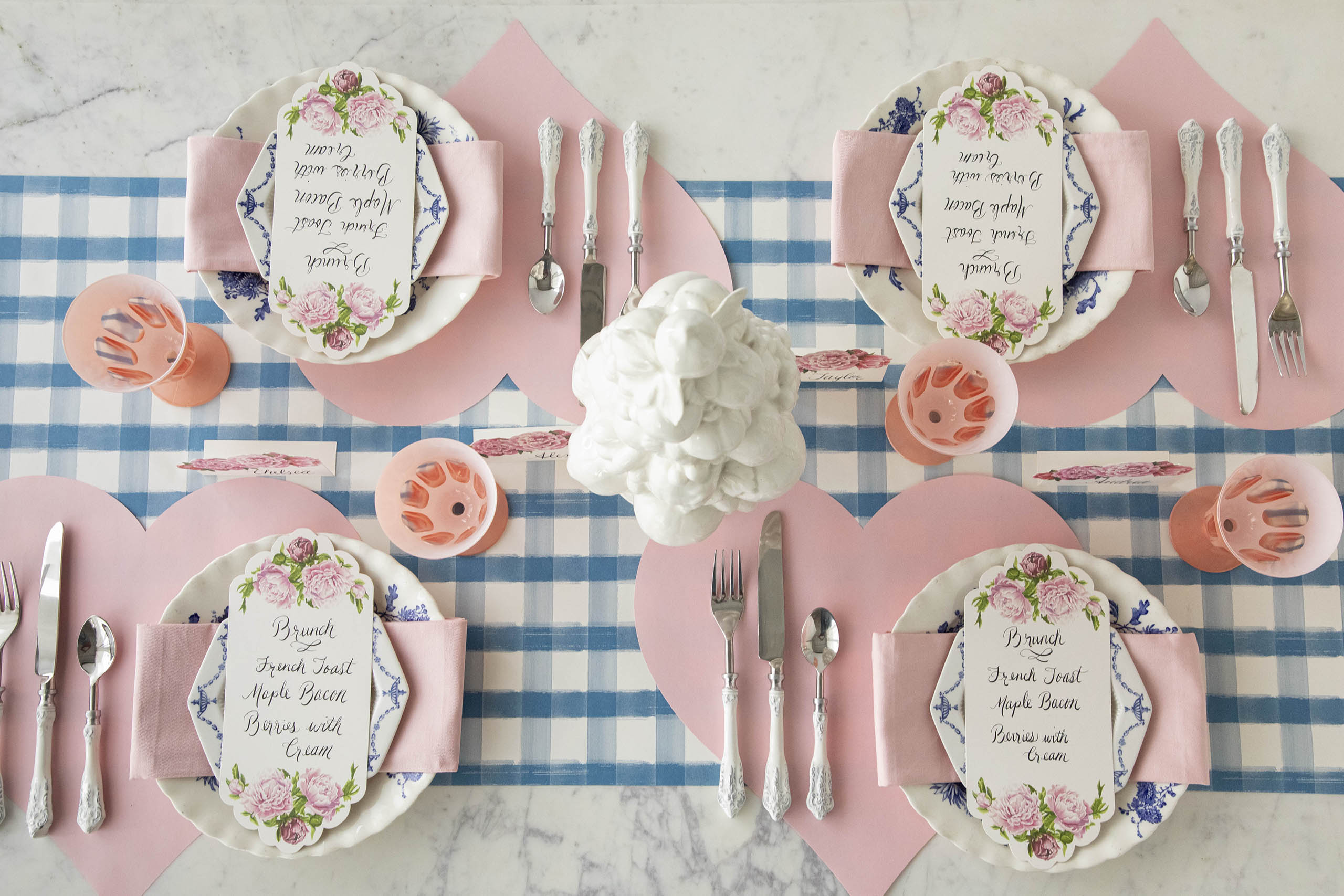 A valentine's table setting featuring Die-cut Pink Heart Placemats, Peony Place Cards and Table Cards, and pink napkins and glasses on the Blue Painted Check Runner.