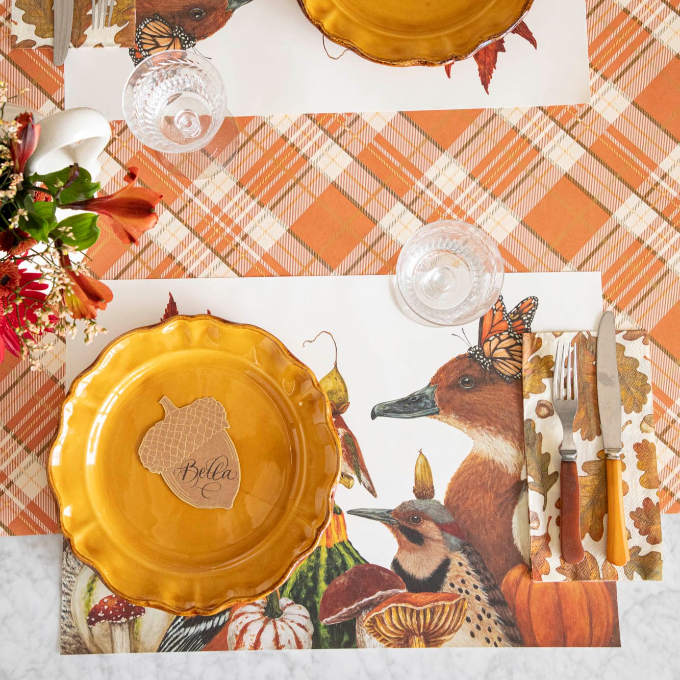 A fall place setting featuring the We Gather Together Placemat, fall leaf napkin, vintage silverware and plate with the Acorn Place Card atop, on the Orange Plaid Runner.