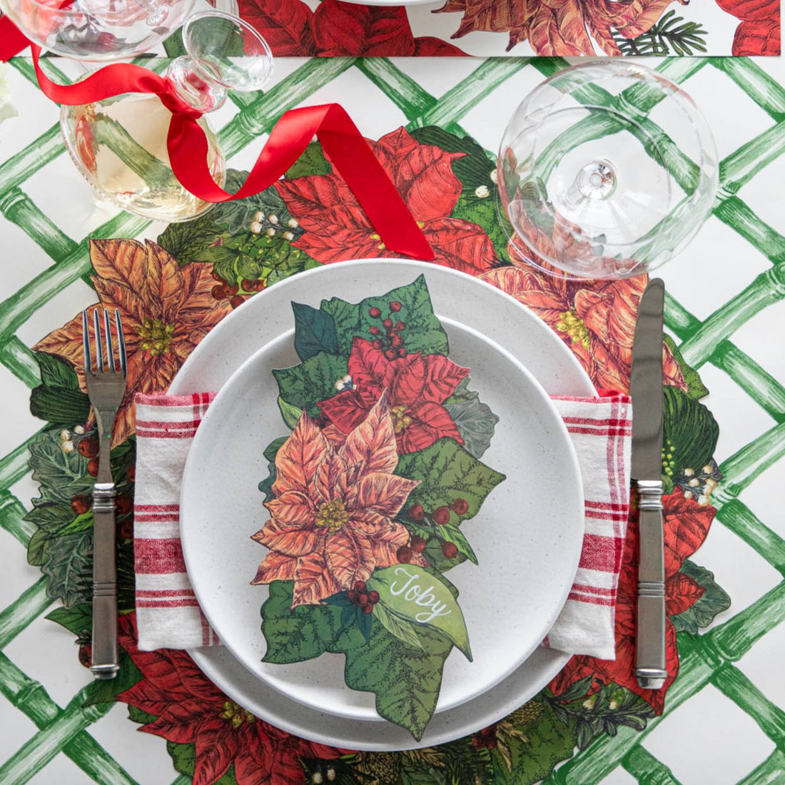 An elegant table setting with the Die-cut Poinsettia Placemat on top of the Green Lattice Runner, with the Poinsettia Table Accent with the name &quot;Toby&quot; written on it, on top of a plate.