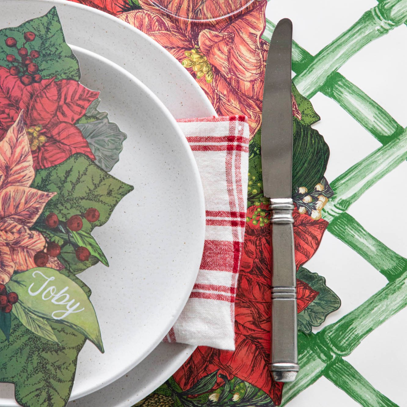 Close up of the Die-cut Poinsettia Wreath Placemat on the Green Lattice Runner with off-white dinner plates and a Poinsettia Table Accent with &quot;Toby&quot; written on the leaf&quot; resting on the plates.