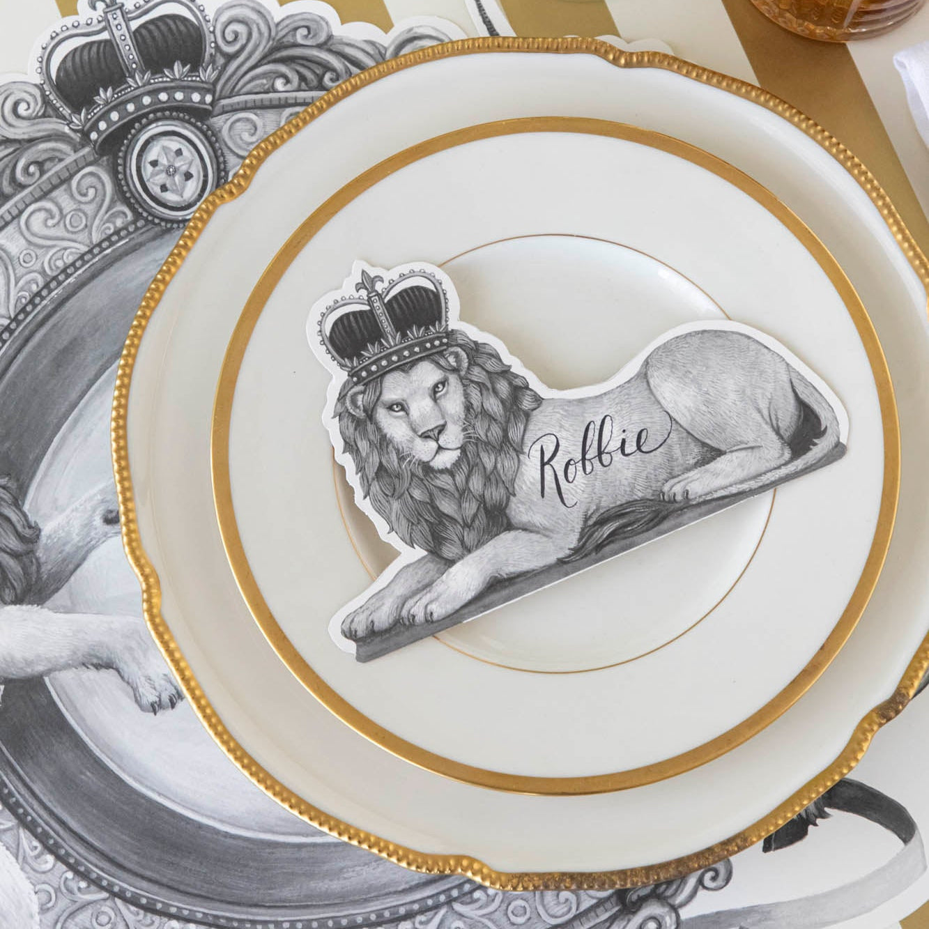 Regal Lion Place Card with &quot;Robbie&quot; written on it, resting on dinner plates on top of the H&amp;C Regal Crest Placemat.