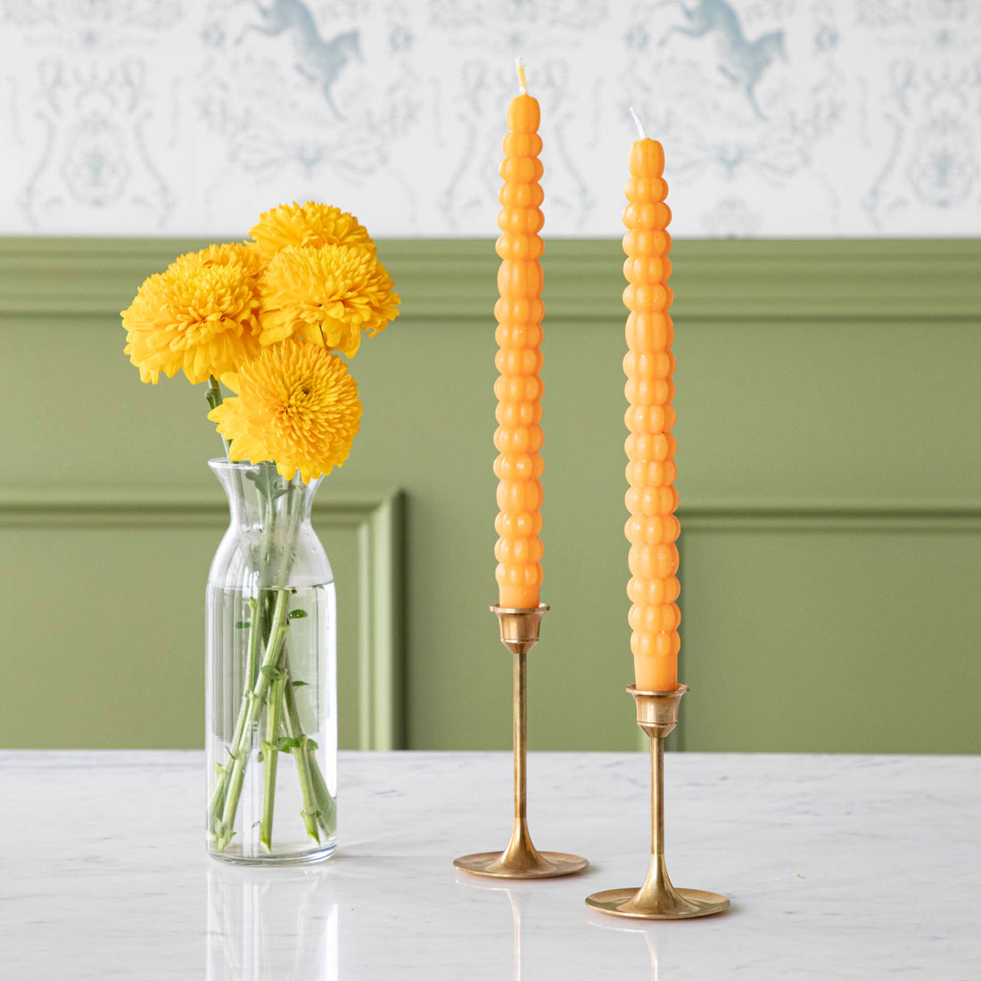 Two orange pumpkin-shaped taper candles on gold-colored candle holders, with a vase of yellow flowers in the background.