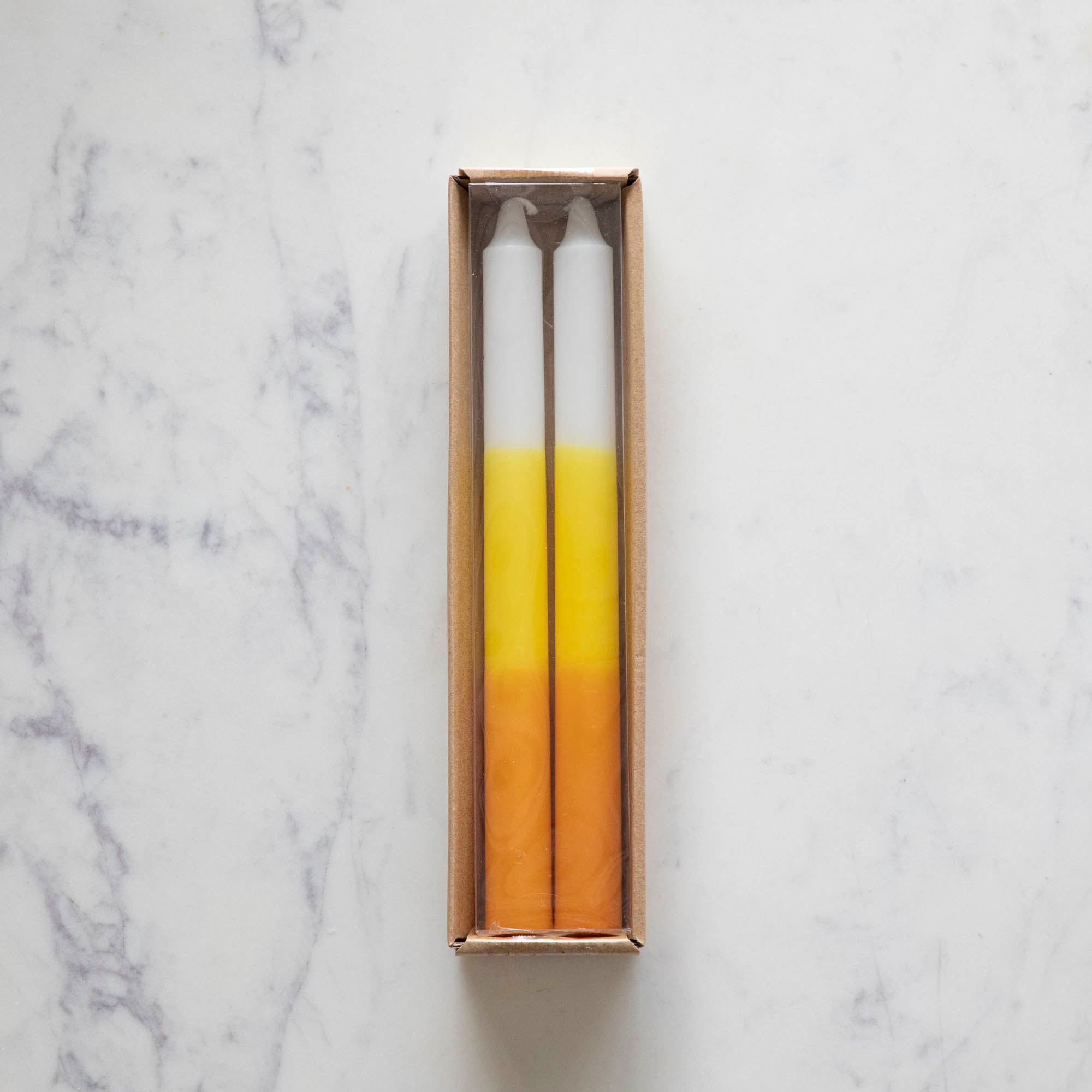 Two taper candles with a yellow, orange, and white color gradient design, resembling candy corn in a box. 