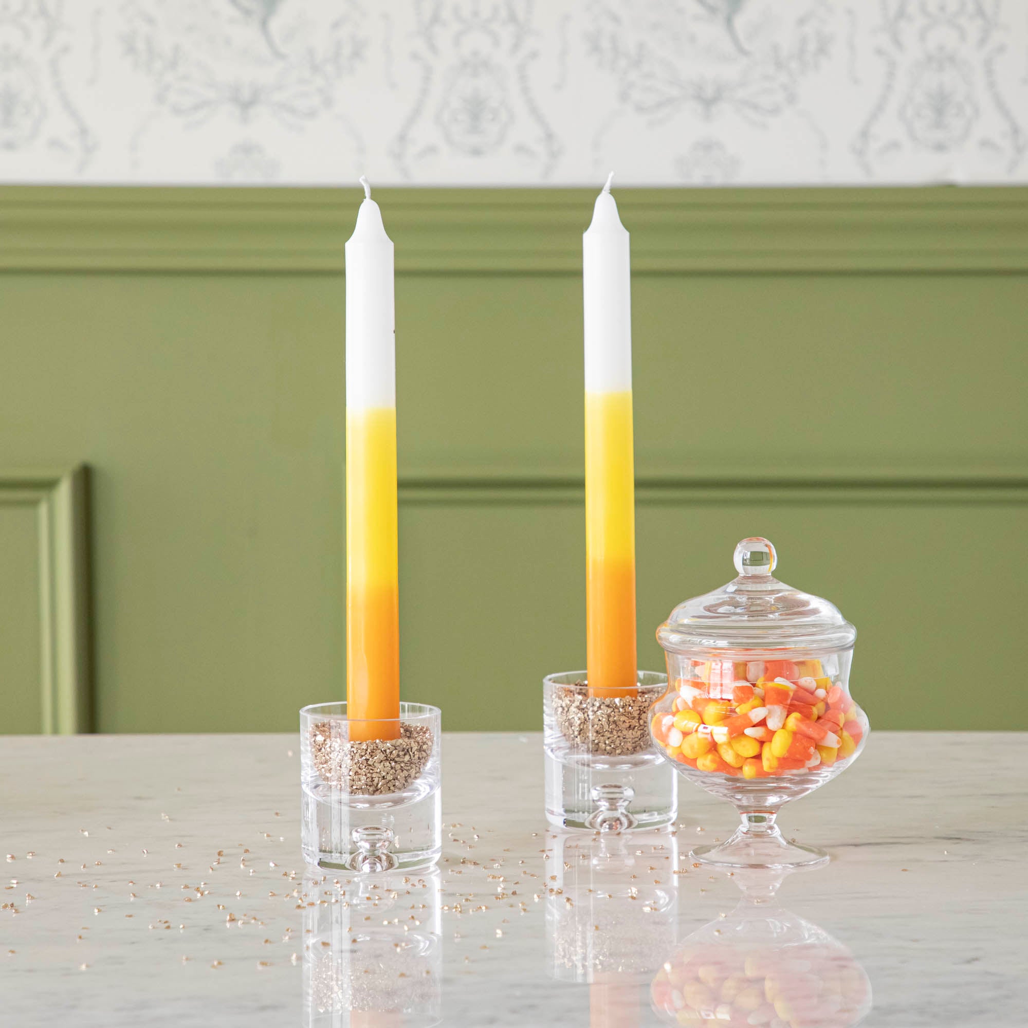 Two taper candles with a yellow, orange, and white color gradient design, resembling candy corn, displayed in front of a green wall.