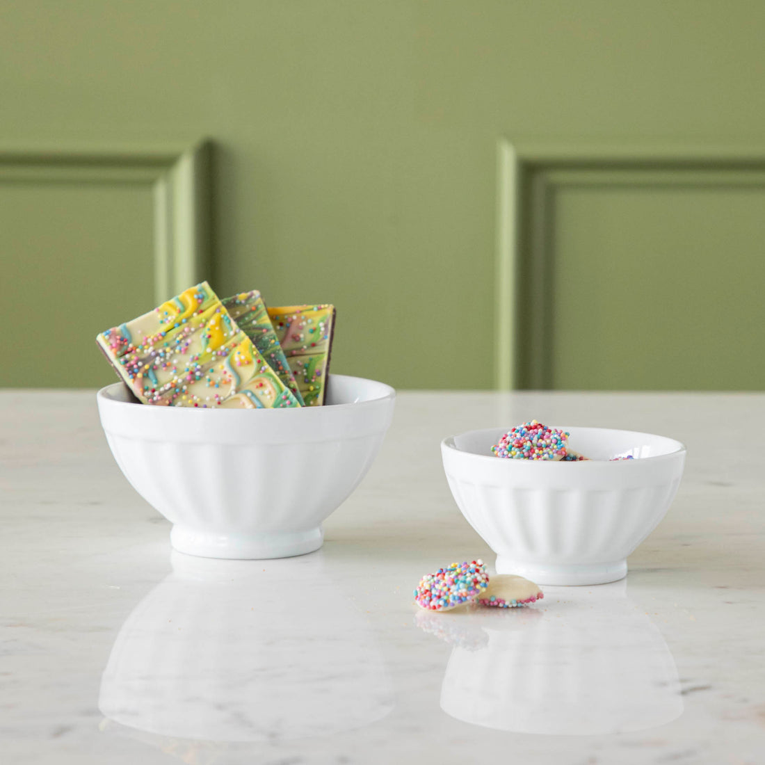 Two white BIA porcelain fluted bowls on a marble surface, one containing sprinkled chocolate bars and the other with chocolate pieces covered in sprinkles.