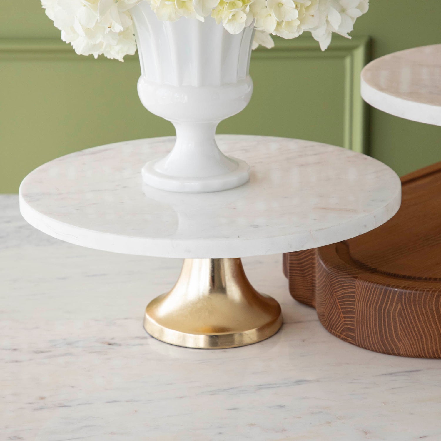 A bouquet of white hydrangeas in a white vase on a Bidk Home Marble Cake Plate with Antique Brass Base with decorative tables and a textured background.