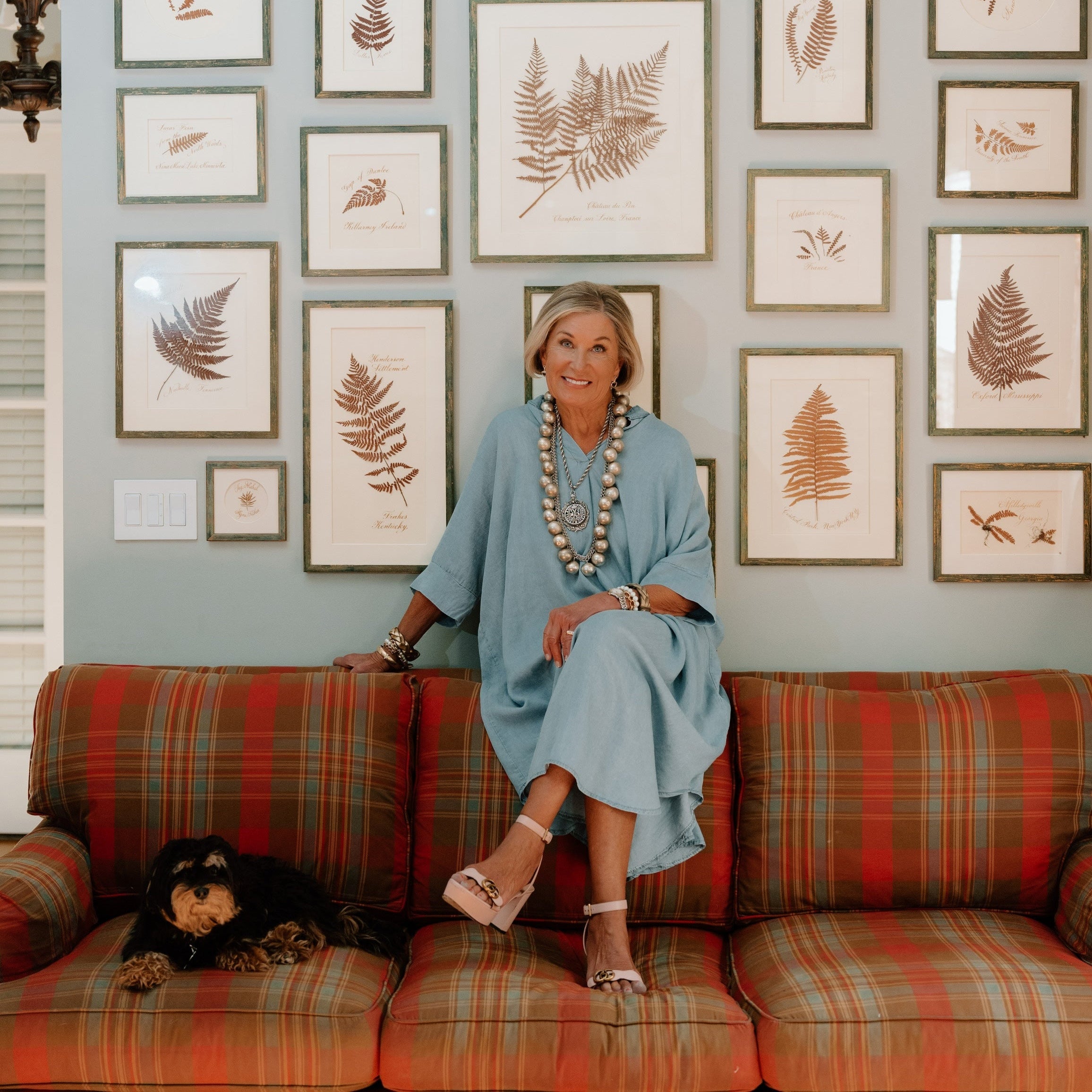 Artist Roiann Ridley in a blue dress sitting on a plaid couch with a wall of framed botanical prints behind her.