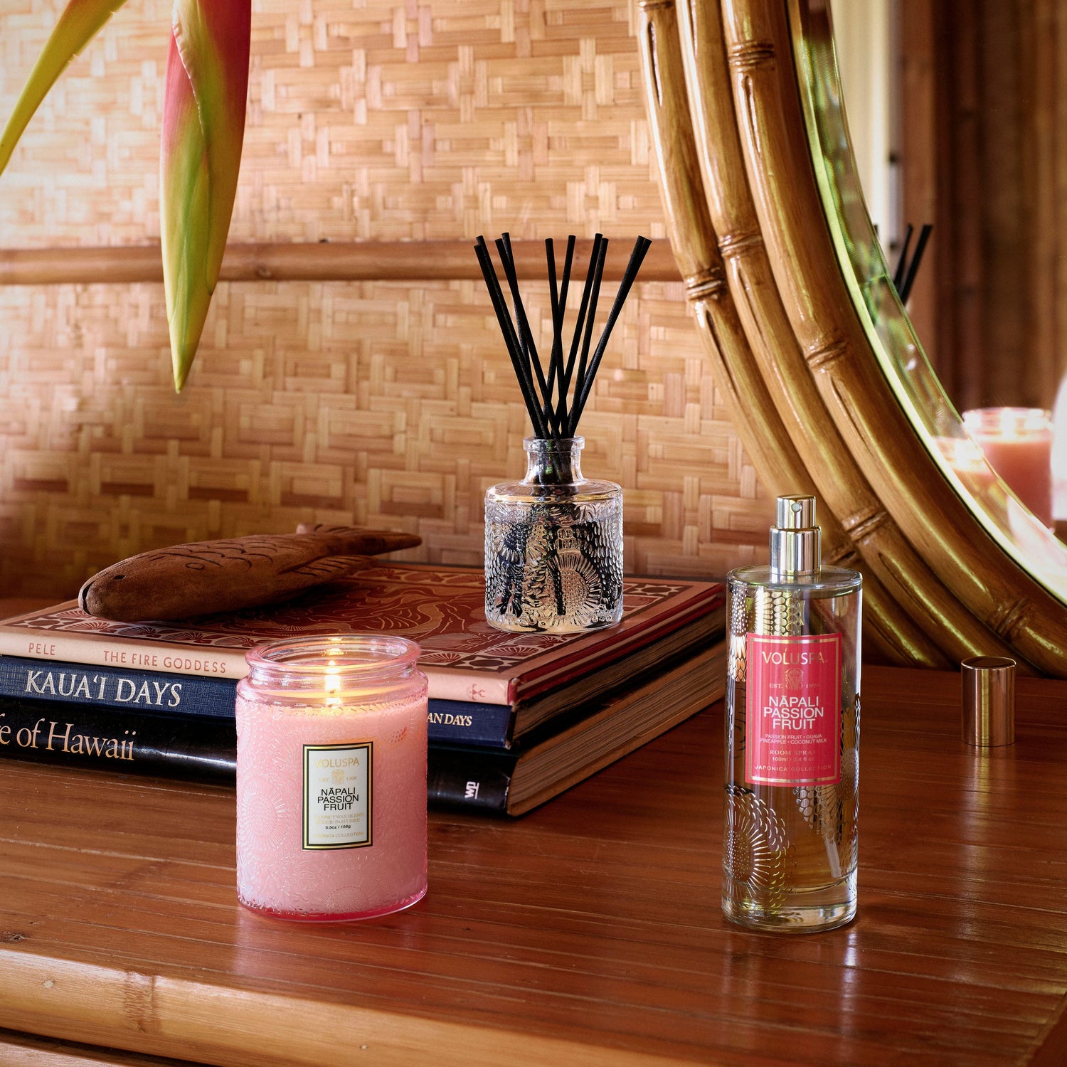 Candle, diffuser, and perfume bottle on a wooden surface with books and a mirror in the background.