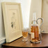 Bottle of rose wine with a glass and a framed drawing on a wooden surface.