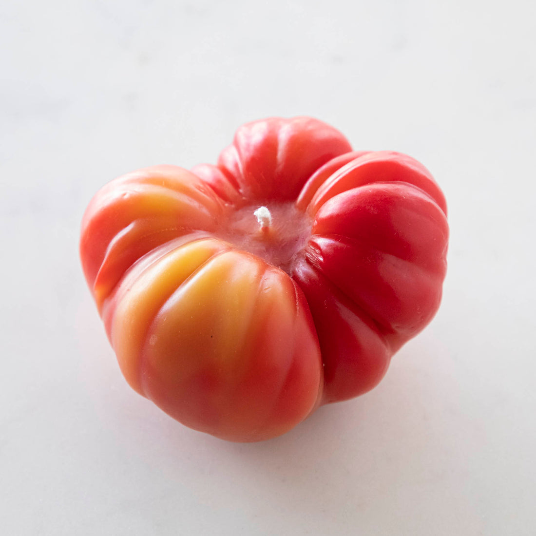 A close-up of an unlit orange-red tomato candle against a white background. 