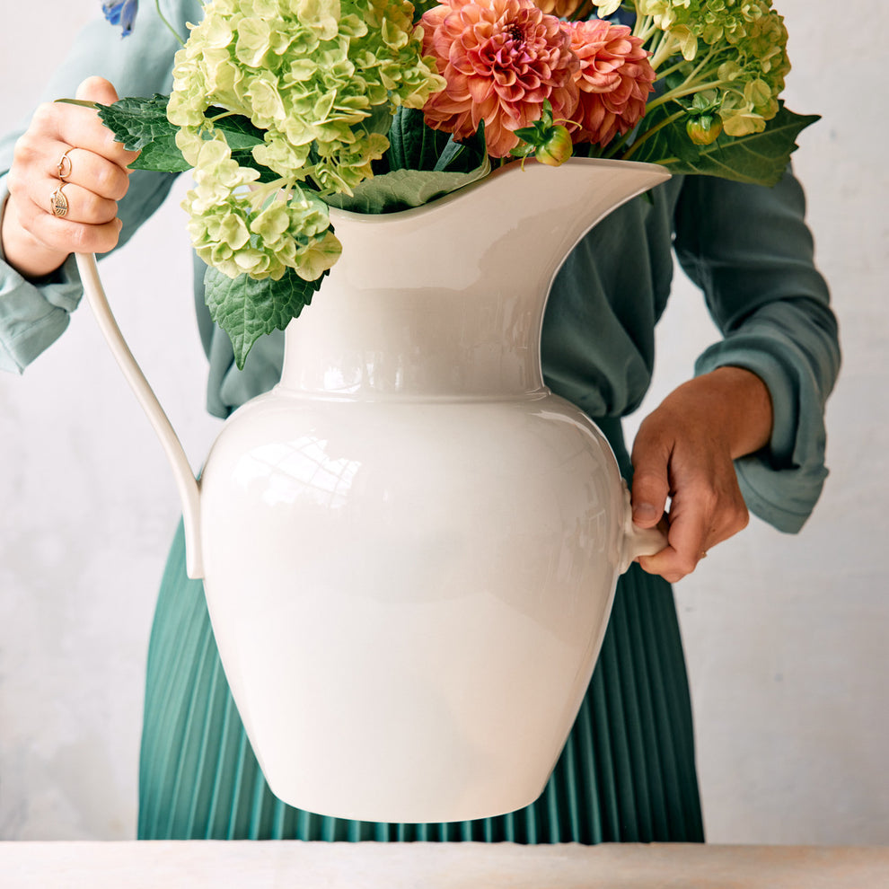 Person holding a white pitcher filled with flowers against a plain background.
