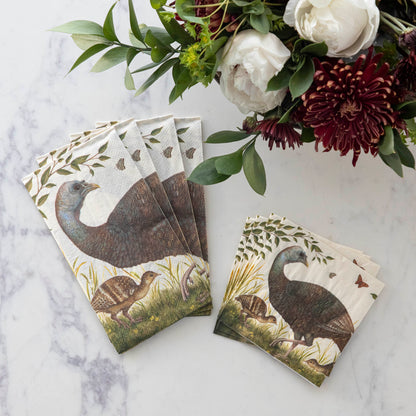 Two stacks of four Heritage Hen Napkins, one Guest and one Cocktail, fanned out on a white table next to a vase of flowers.