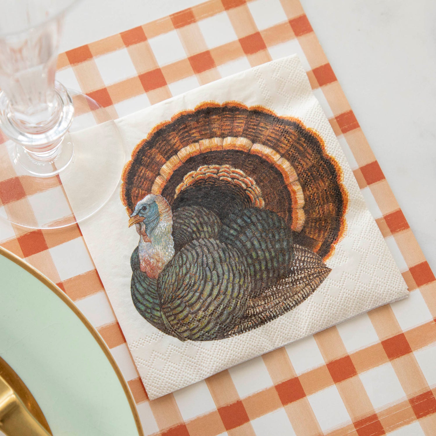 A Heritage Turkey Cocktail Napkin under a stemmed glass in a Thanksgiving place setting.