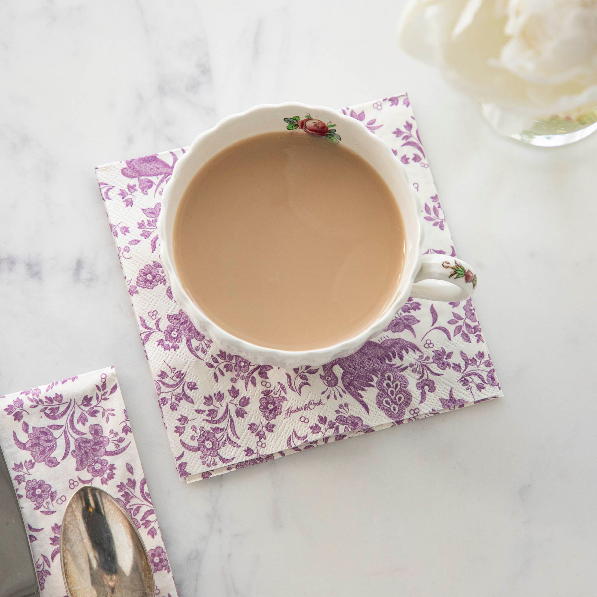 A cup of coffee sits on a table next to a Lilac Regal Peacock Napkin by Hester &amp; Cook.