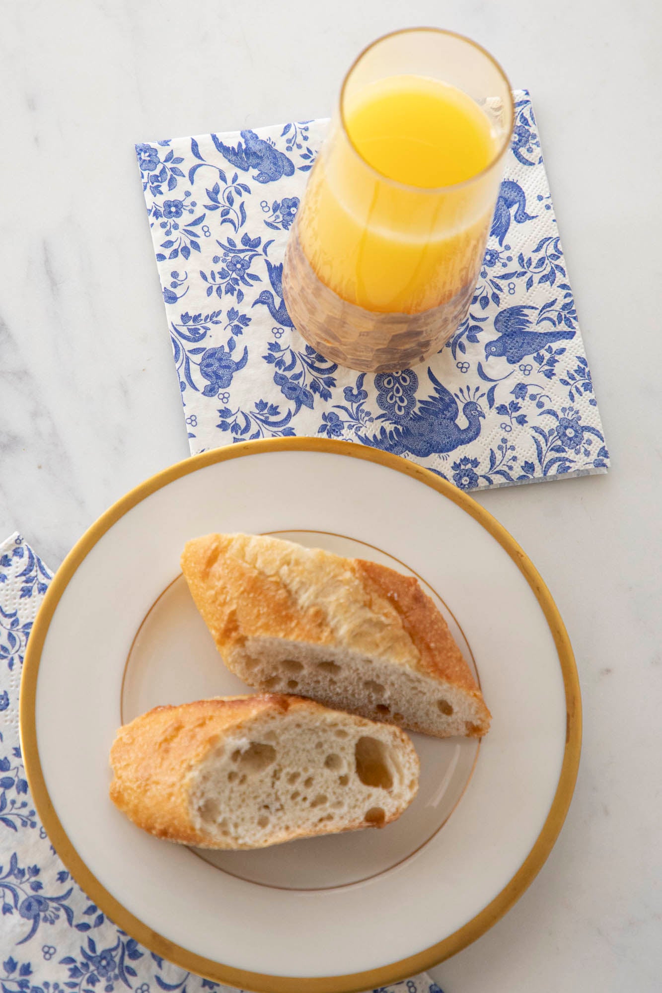 A plate of bread and a glass of orange juice, accompanied by Blue Regal Peacock Napkins from Hester &amp; Cook and decorated with a bird pattern.