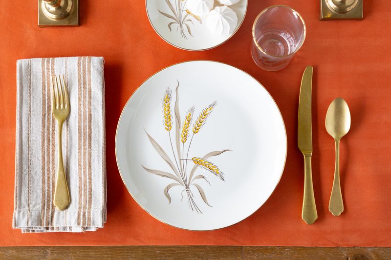 Table setting with white plate, gold cutlery, and napkin on an orange felted table runner.
