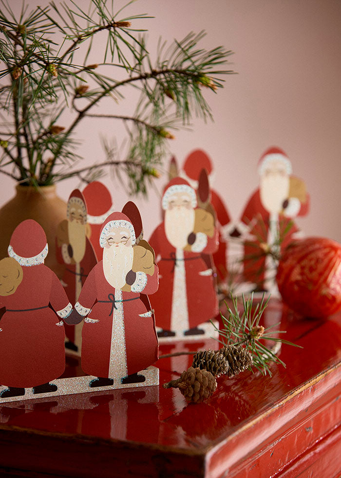 Set of paper Santa Claus paper cut-outs on a reflective surface with Christmas decorations.