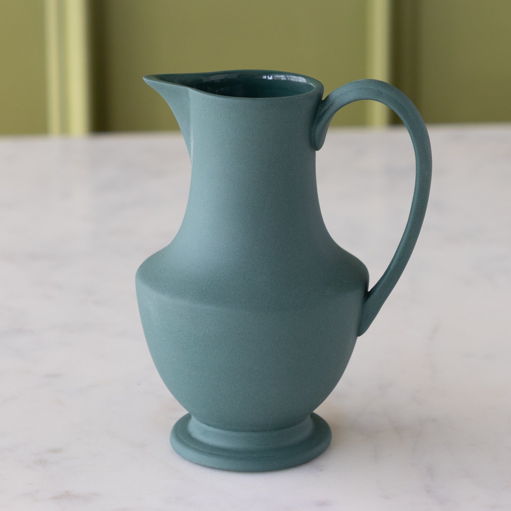 Teal ceramic pitcher on a marble surface with a green background.