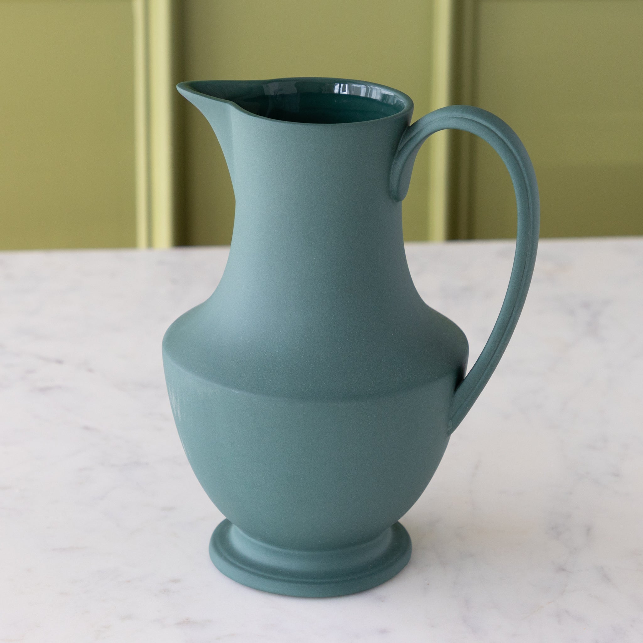 Teal ceramic pitcher on a marble surface with a green background.