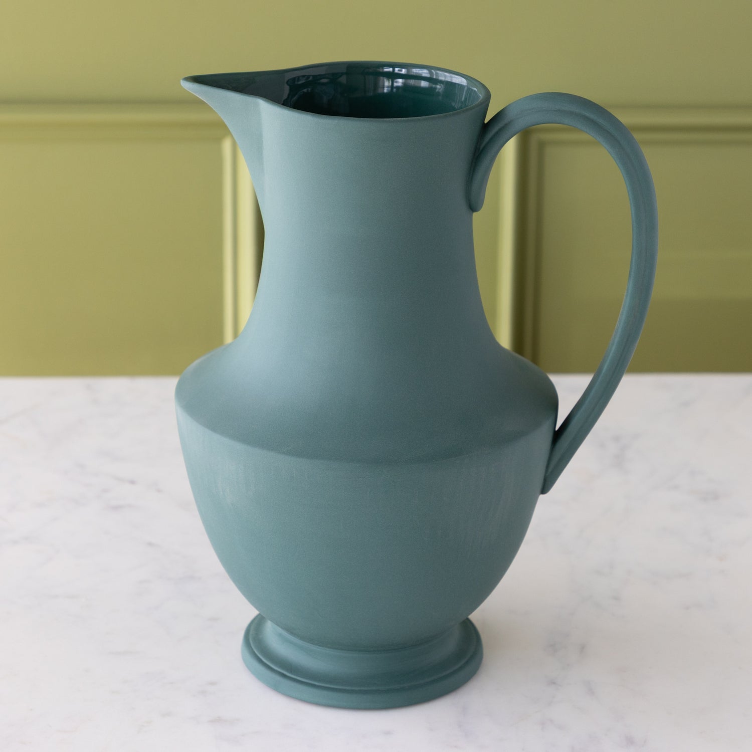 Teal ceramic pitcher on a marble surface with a green background.
