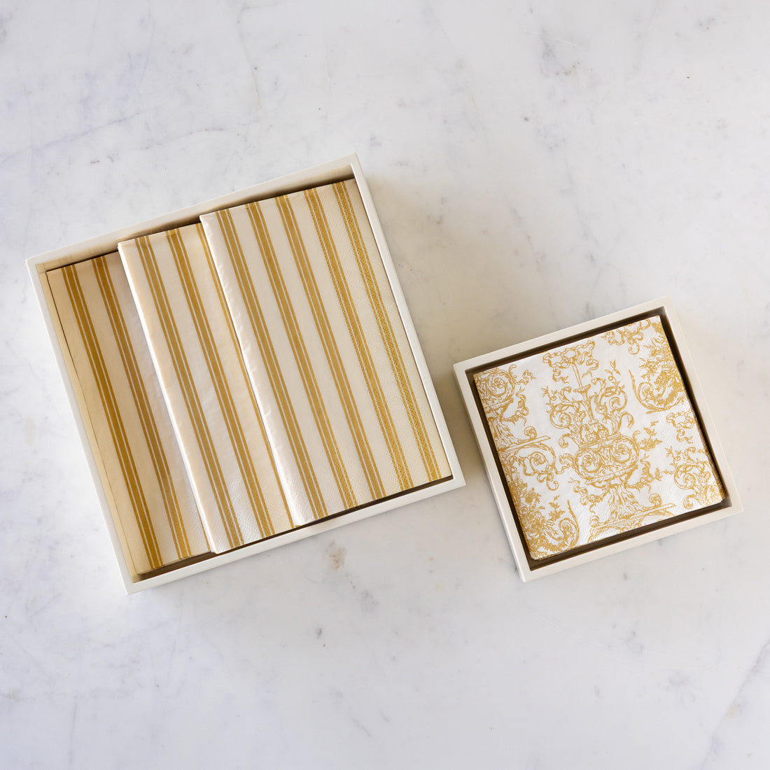 A set of square decorative coasters or tiles in a white box, shown open on a light marble surface. The coasters feature a white and gold striped pattern, and the lid displays a matching gold damask design on white.