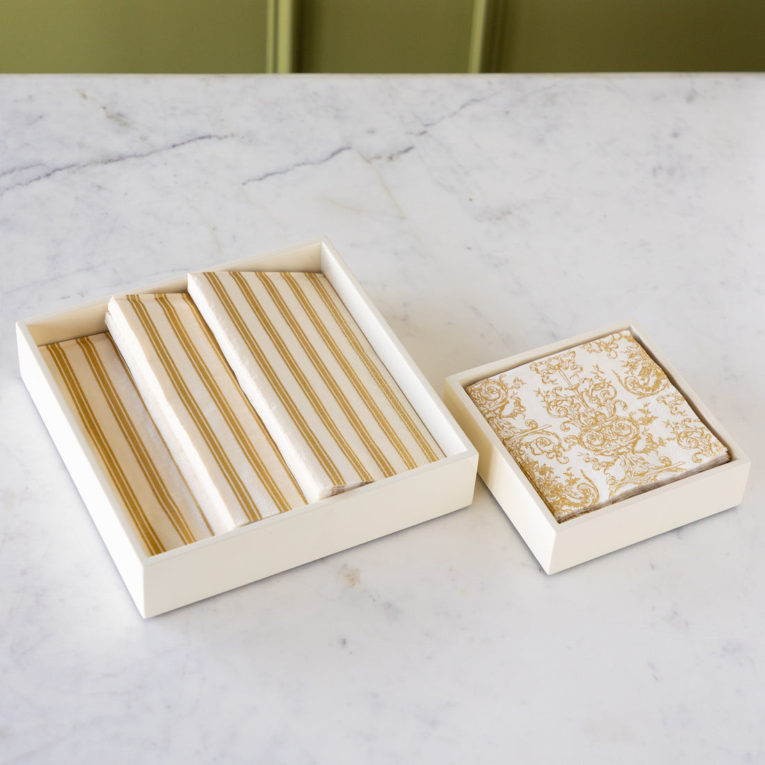 A set of cream and gold striped napkins displayed in an open cream linen box next to a matching square box lid with a gold damask pattern, set on a white marble countertop.