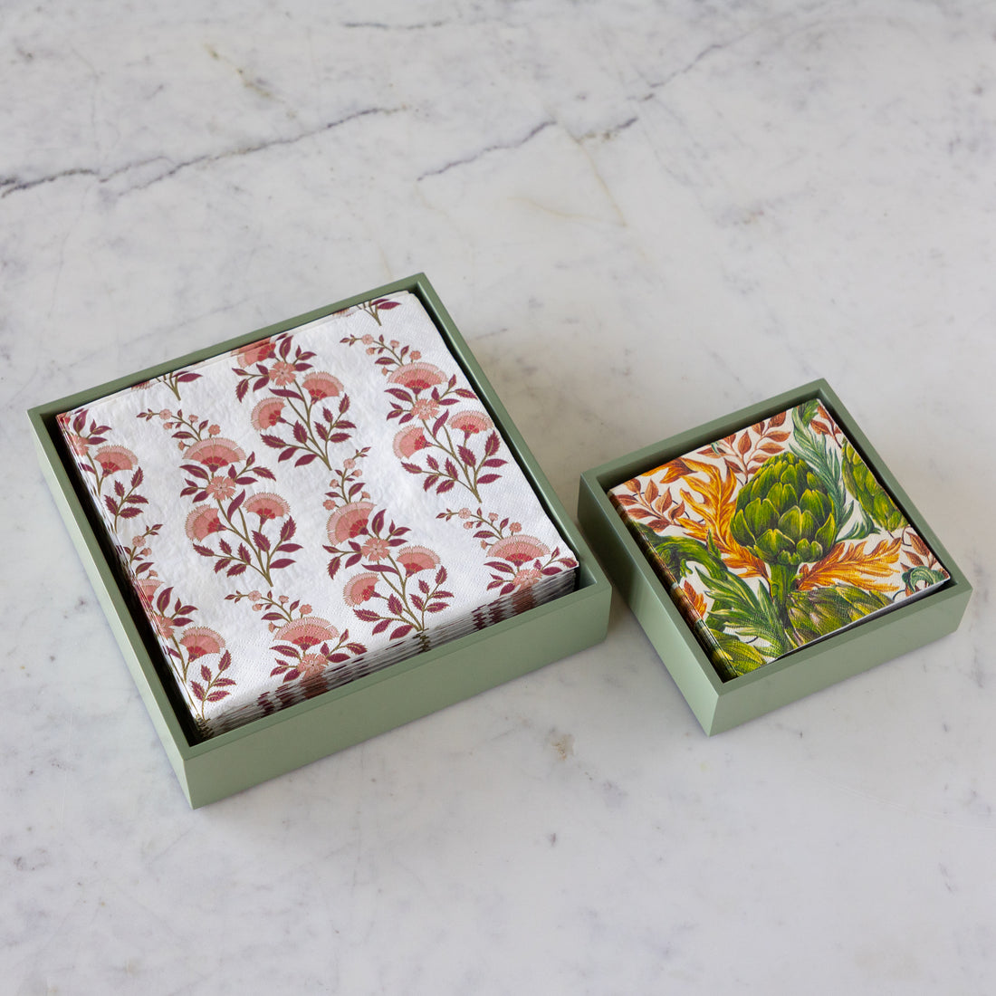 Two square decorative boxes on a light marble surface, one open to show a white napkin with a pink and brown floral pattern and one closed showing a colorful floral-and-vegetable print on the lid.