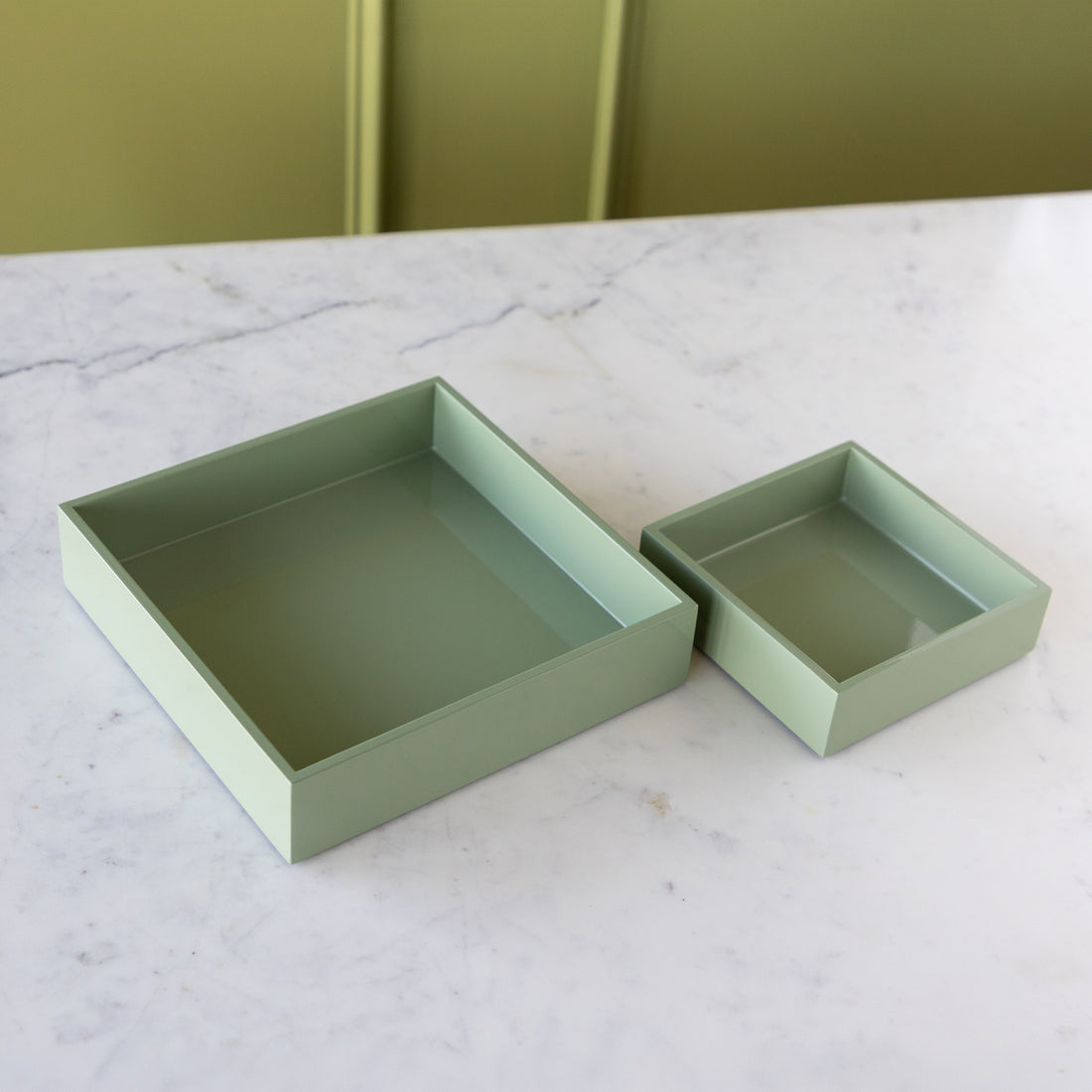 Two square shallow boxes in a muted sage green color, one larger and one smaller, displayed on a white marble surface with a green paneled background.