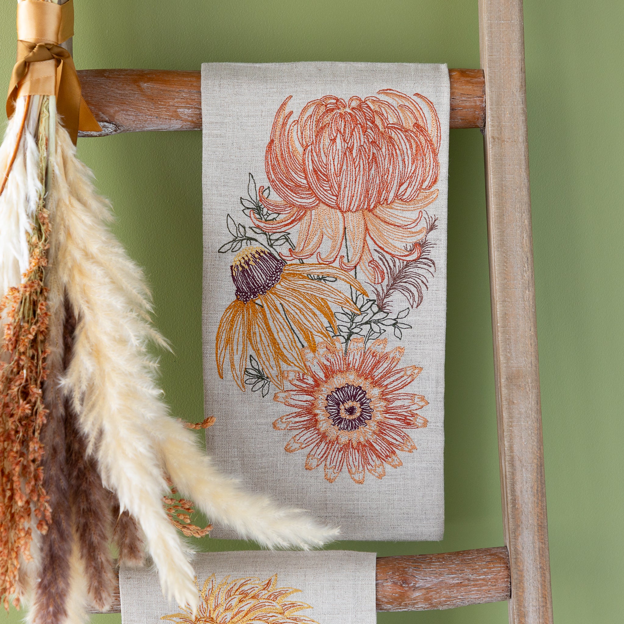 Autumn Mums Tea Towel hanging on a wooden ladder against a green wall.