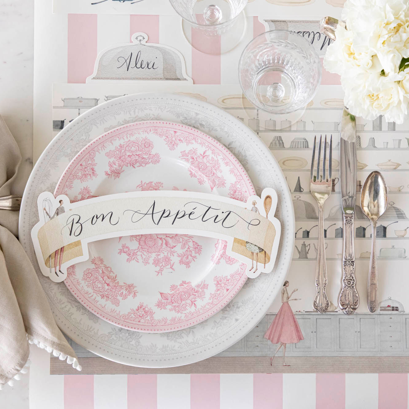 Two Cooks Banner Table Accent with &quot;Bon Appetit&quot; written on it, resting on dinner plates, on an elegant place setting for one.