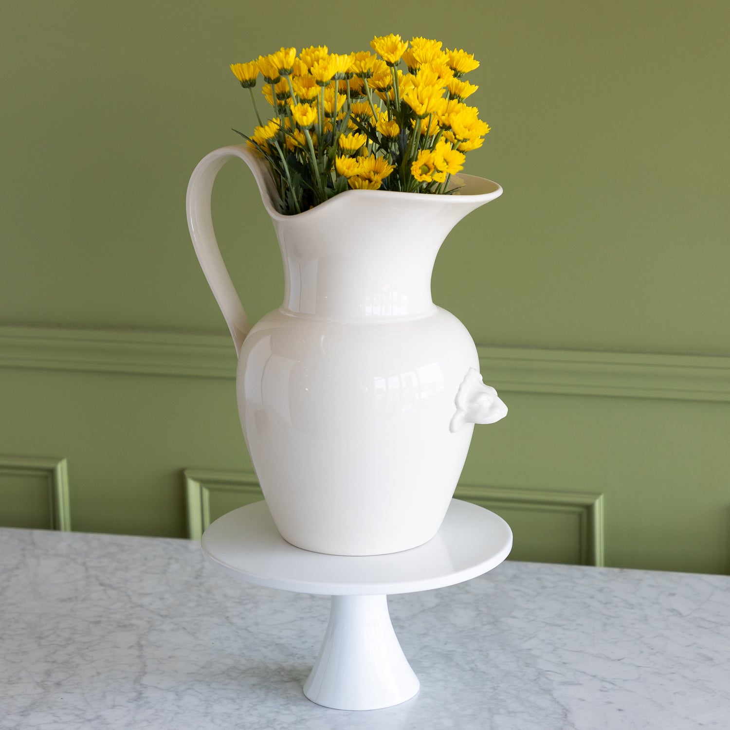 White pitcher with yellow flowers on a marble surface against a green wall.