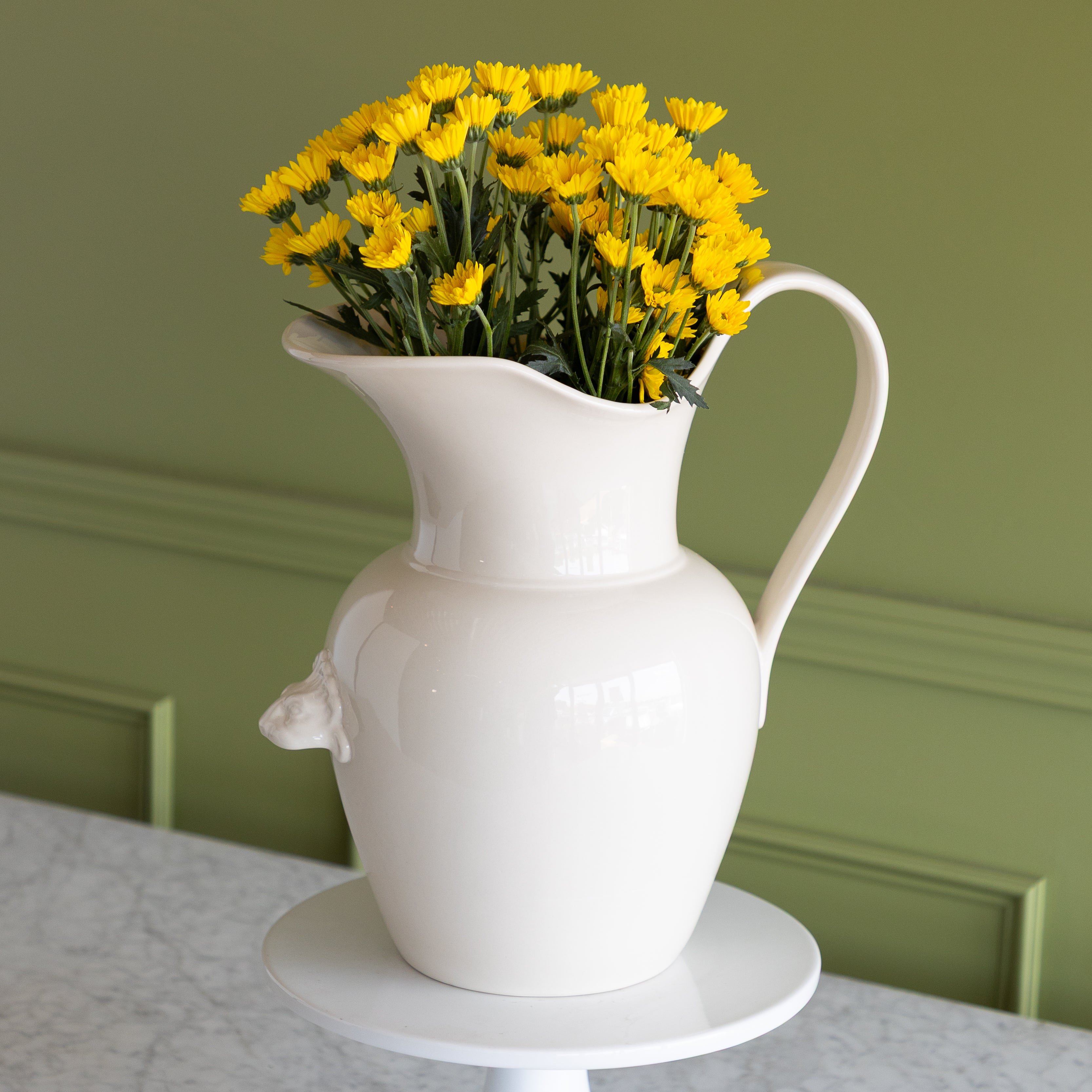 White pitcher with yellow flowers on a table against a green wall.