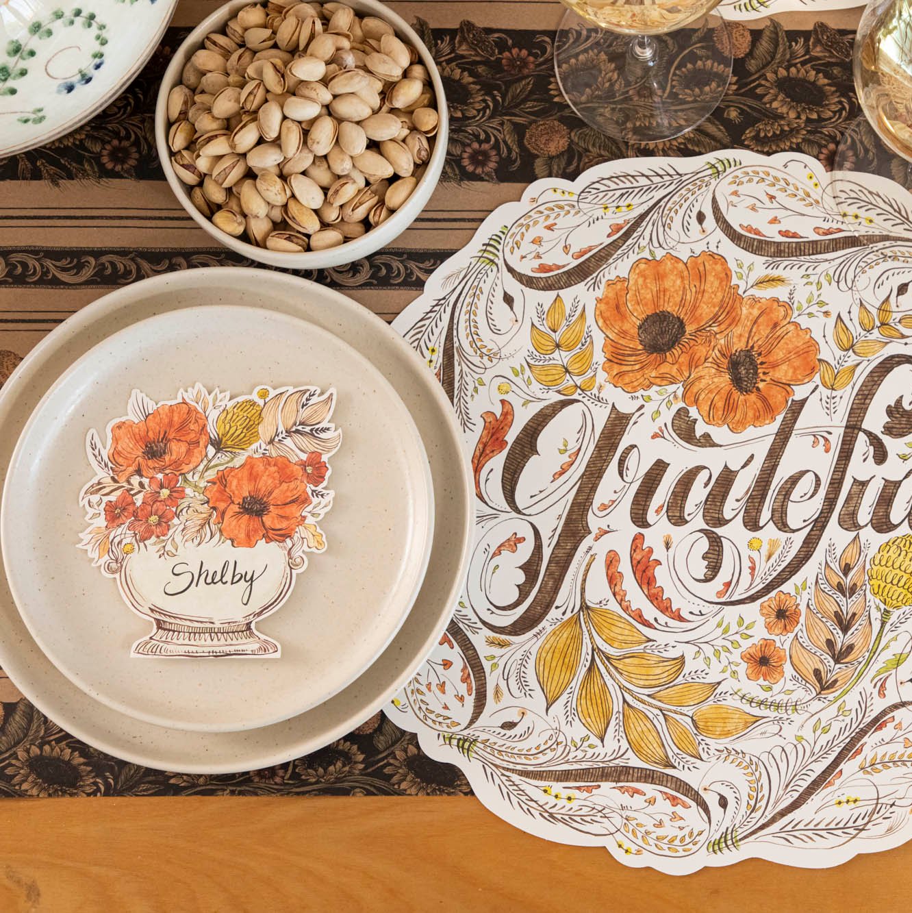 The Autumn Arrangement Place Card with the name "Shelby" written on it, on a fall themed table setting with a floral placemat with "Grateful" written on it.