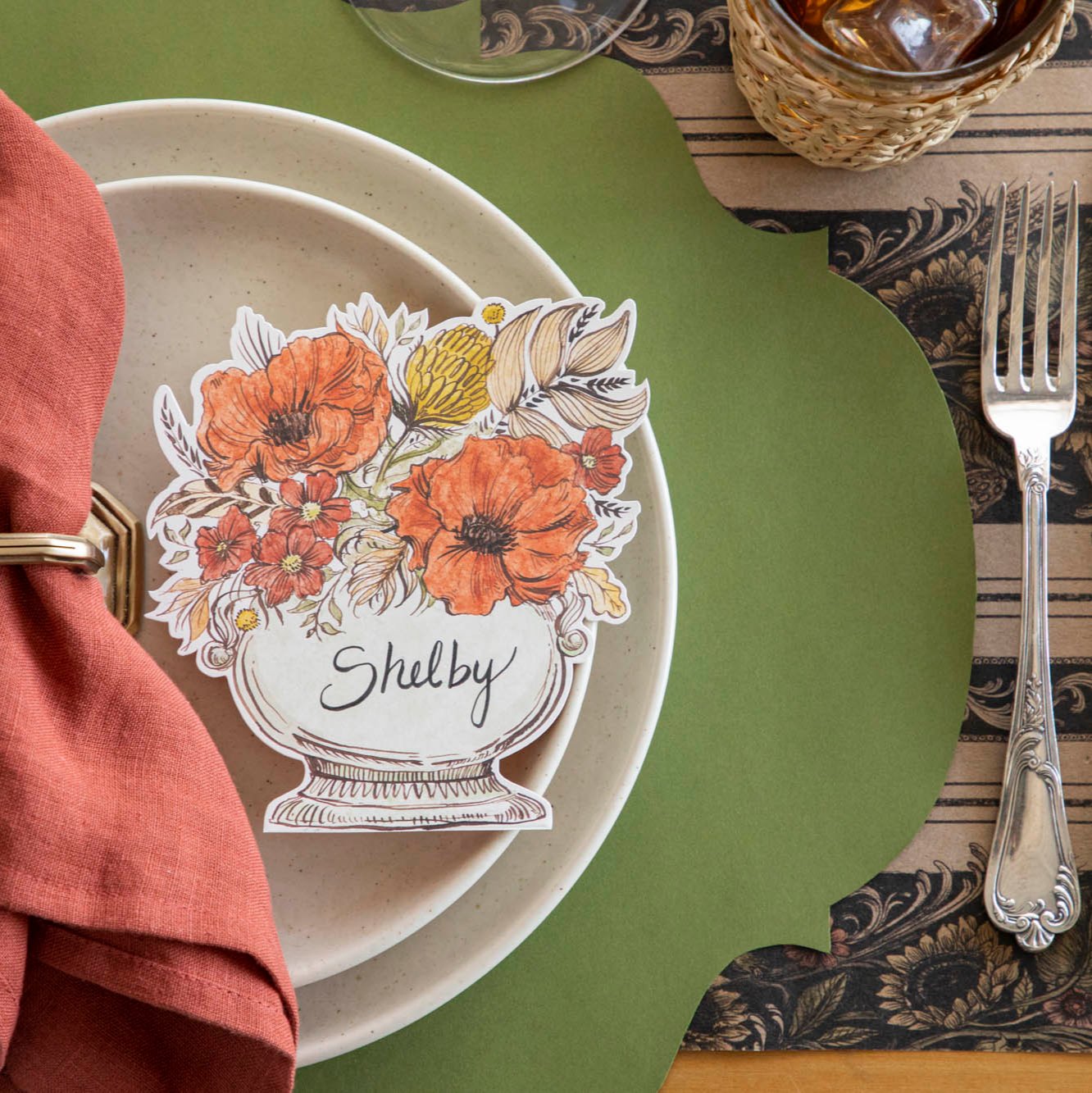 The Autumn Arrangement Place card with the name "Shelby" written on it, on top of salt dinner plates, a Green French Frame placemat and Vintage Sunflower Stripe runner underneath.
