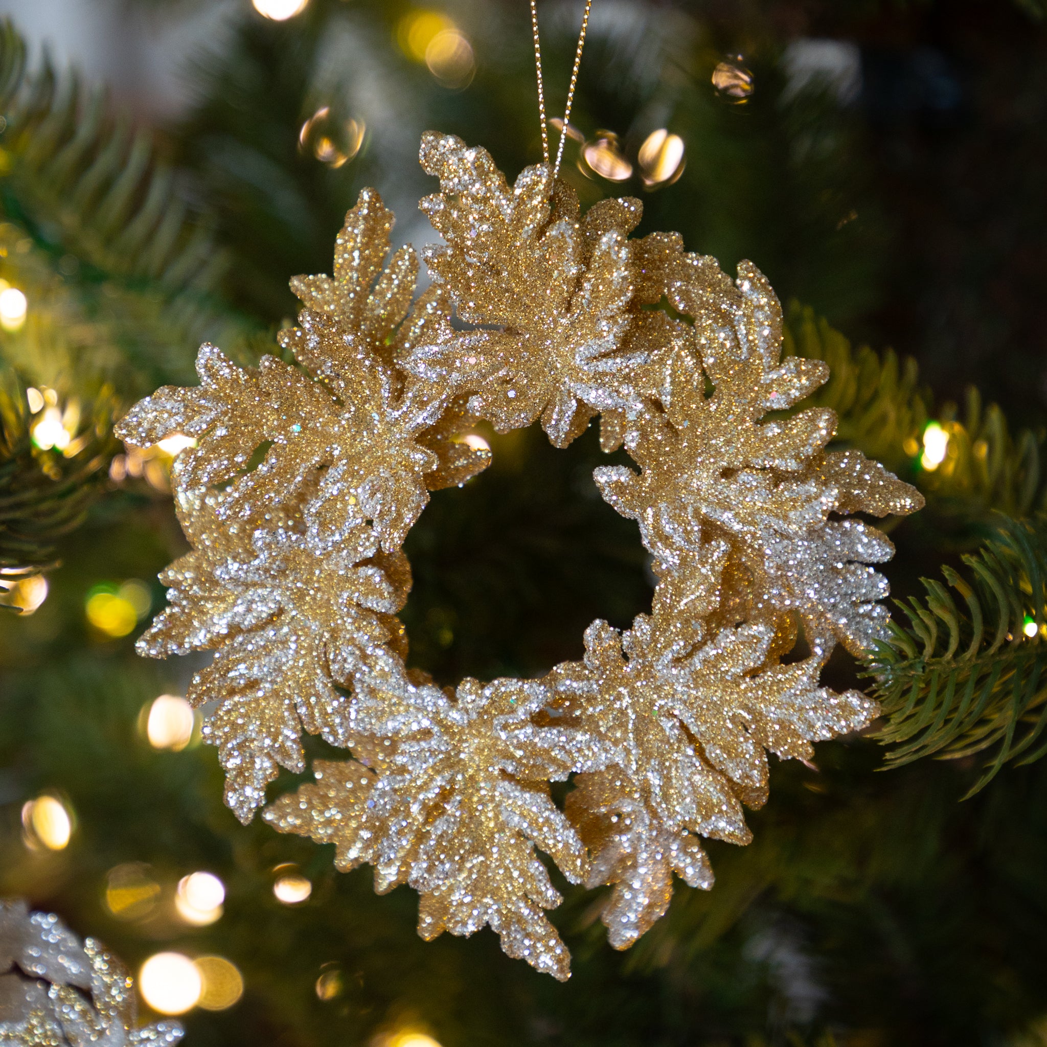 Gold glittery wreath ornament on a Christmas tree with blurred lights in the background.