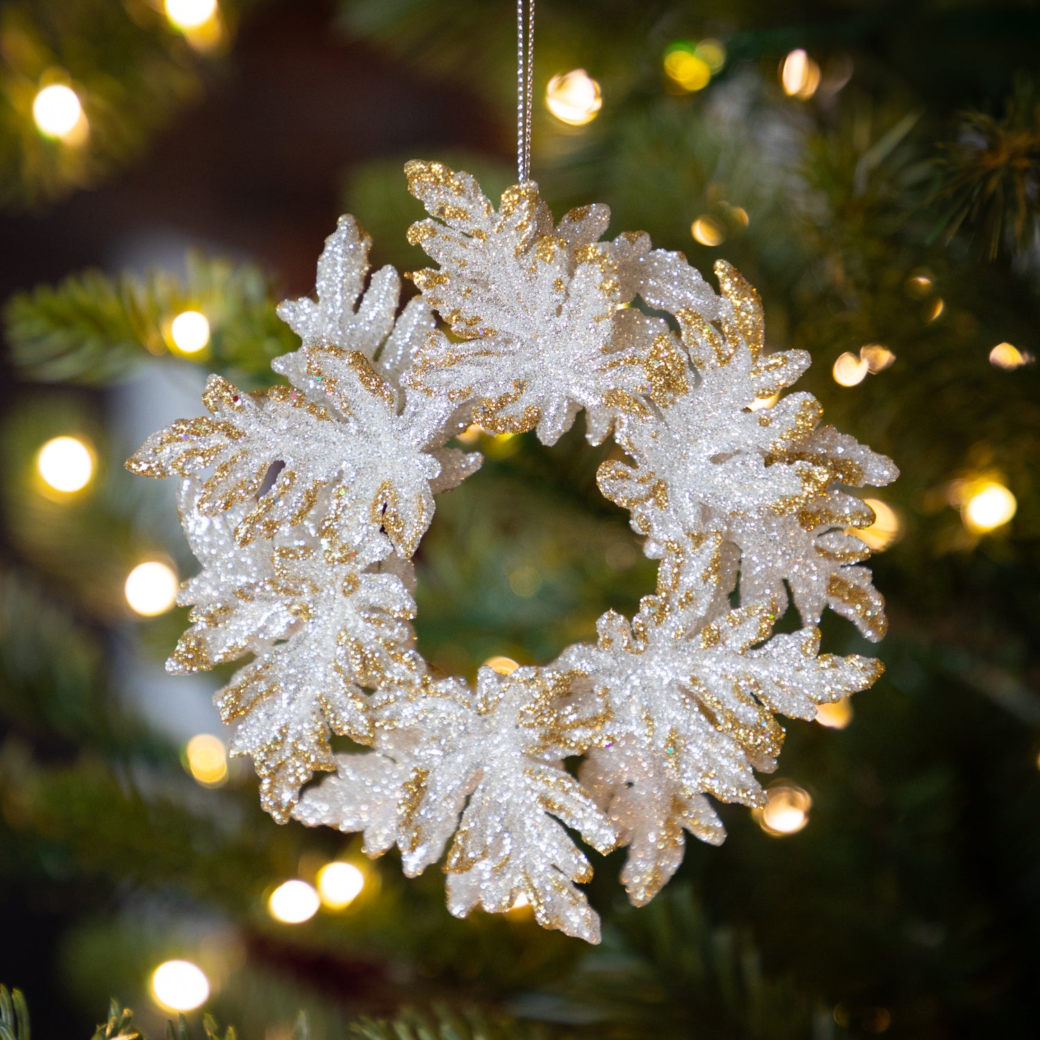 Decorative silver wreath with gold tips ornament on a Christmas tree with blurred lights in the background.