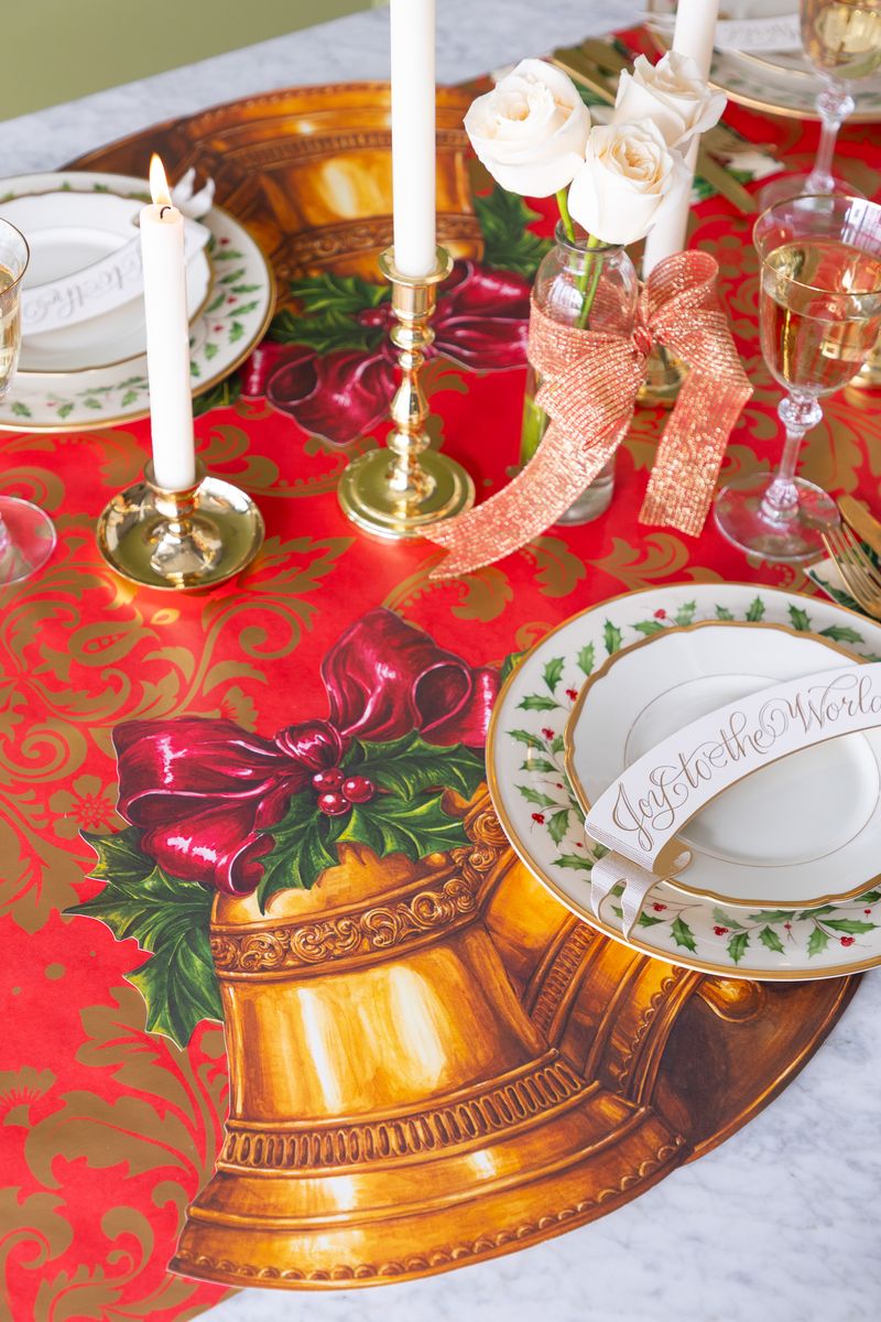 Christmas-themed table setting with red and gold table runner, gold bell paper placemat, and white candles.