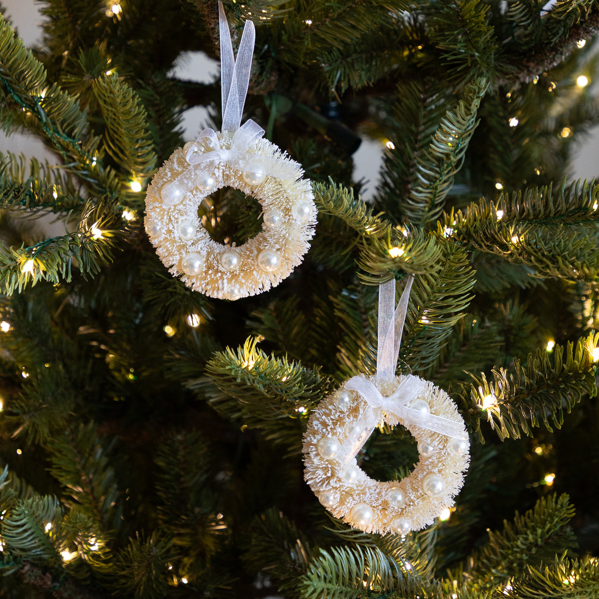 Two round Christmas wreath ornaments with pearl-like beads and white ribbon hangers, hanging on a green Christmas tree with warm string lights.