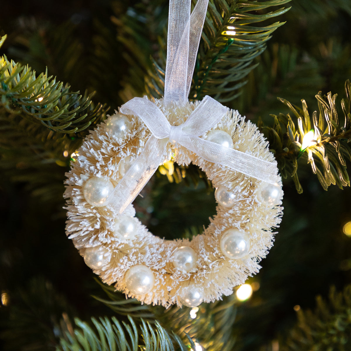 A frosted white Christmas wreath ornament decorated with white pearl-like beads and a sheer white ribbon bow, hanging on a green Christmas tree with warm string lights.