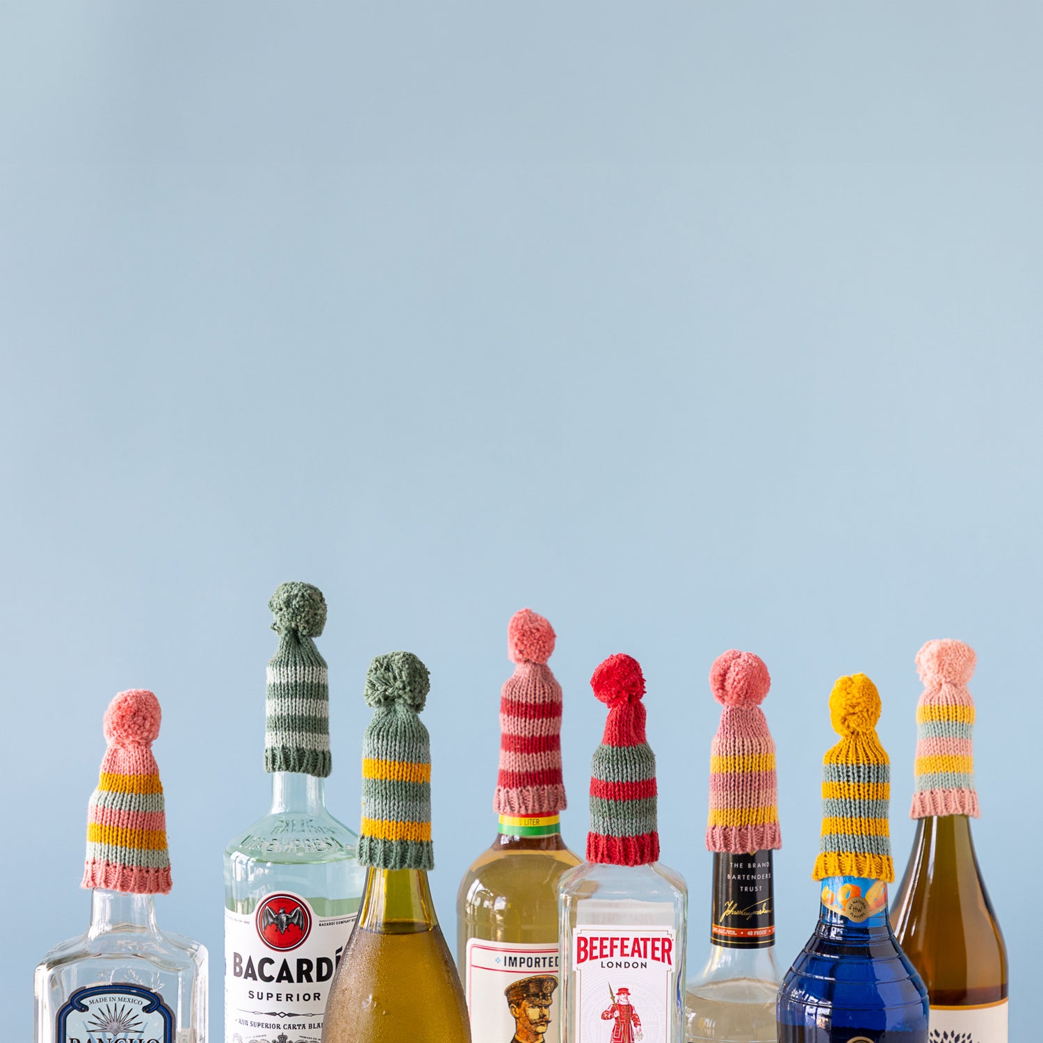 Collection of alcohol bottles with colorful knit hats on a light blue background.