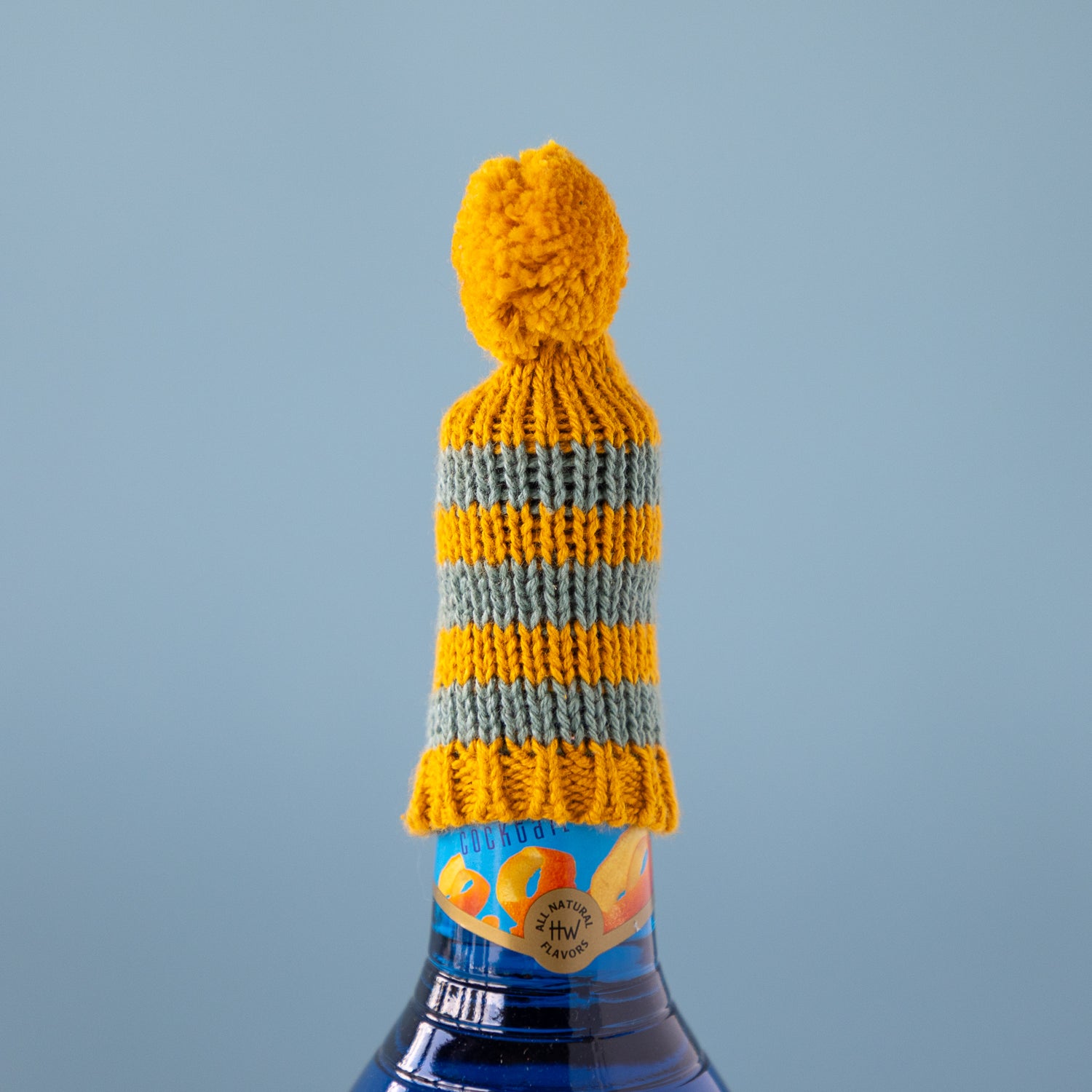 Knitted bottle cover with yellow and green stripes on a blue bottle against a light blue background.